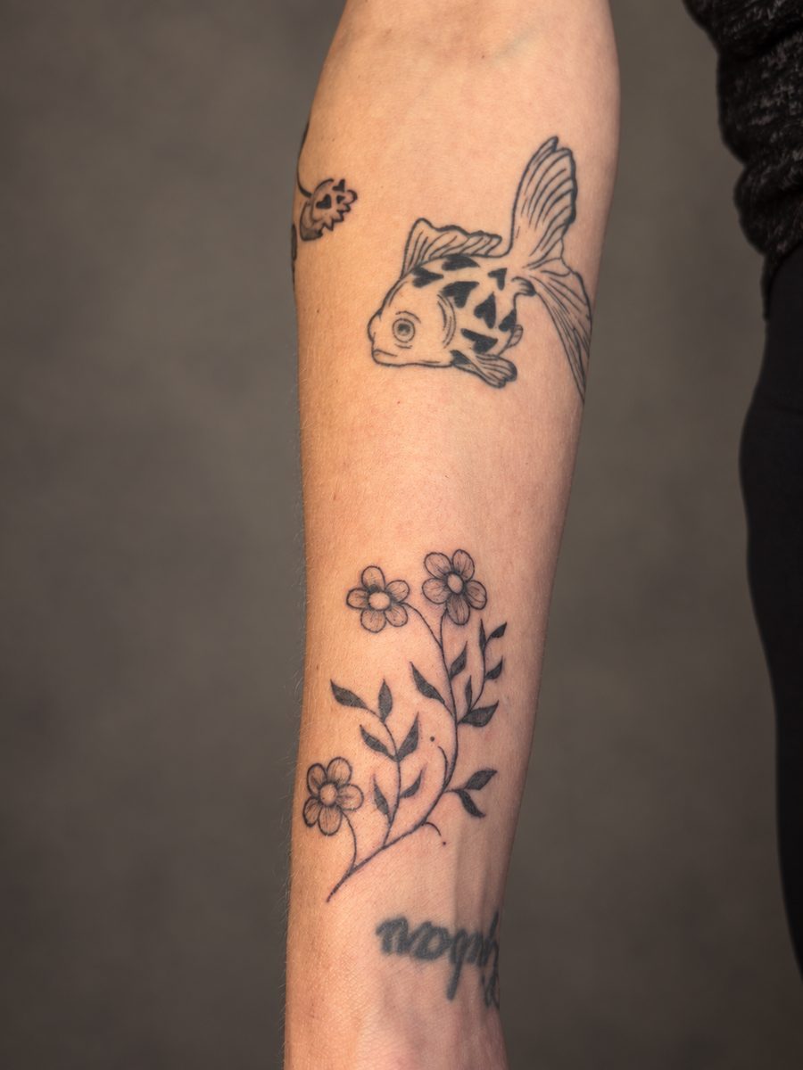 Fineline tattoo van een vis met kleine bloemen op de onderarm, gezet in Lelystad